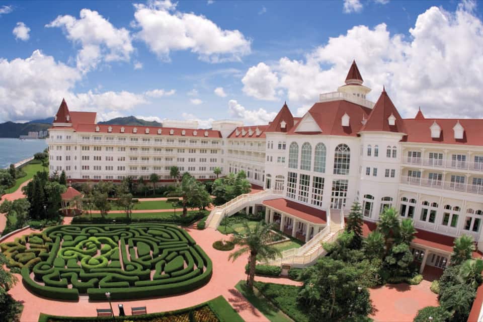 Um edifício grande, branco, com vários andares e telhados vermelhos, com um jardim paisagístico e um labirinto.