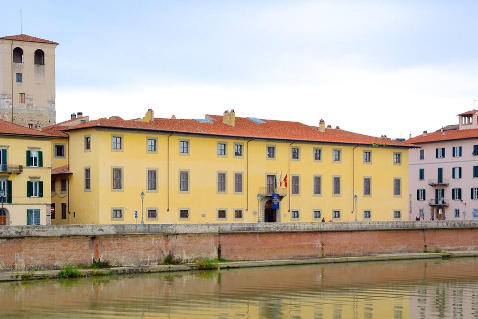 A canal with buildings along the banks, including a yellow building with a tower and a pink building with a balcony.