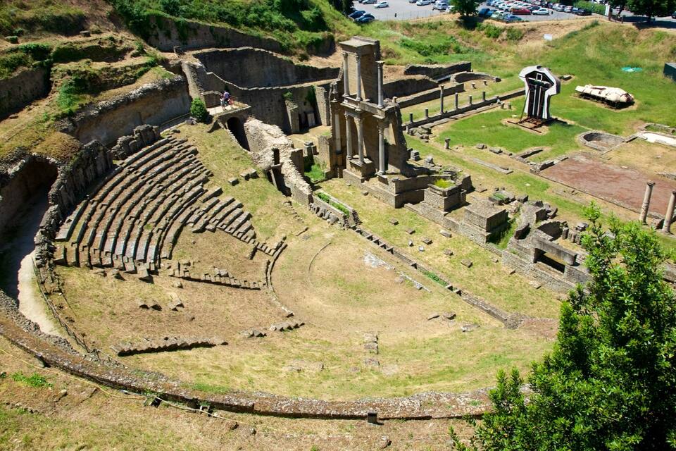 An ancient amphitheater with stone seating and columns, surrounded by grassy areas and ruins.