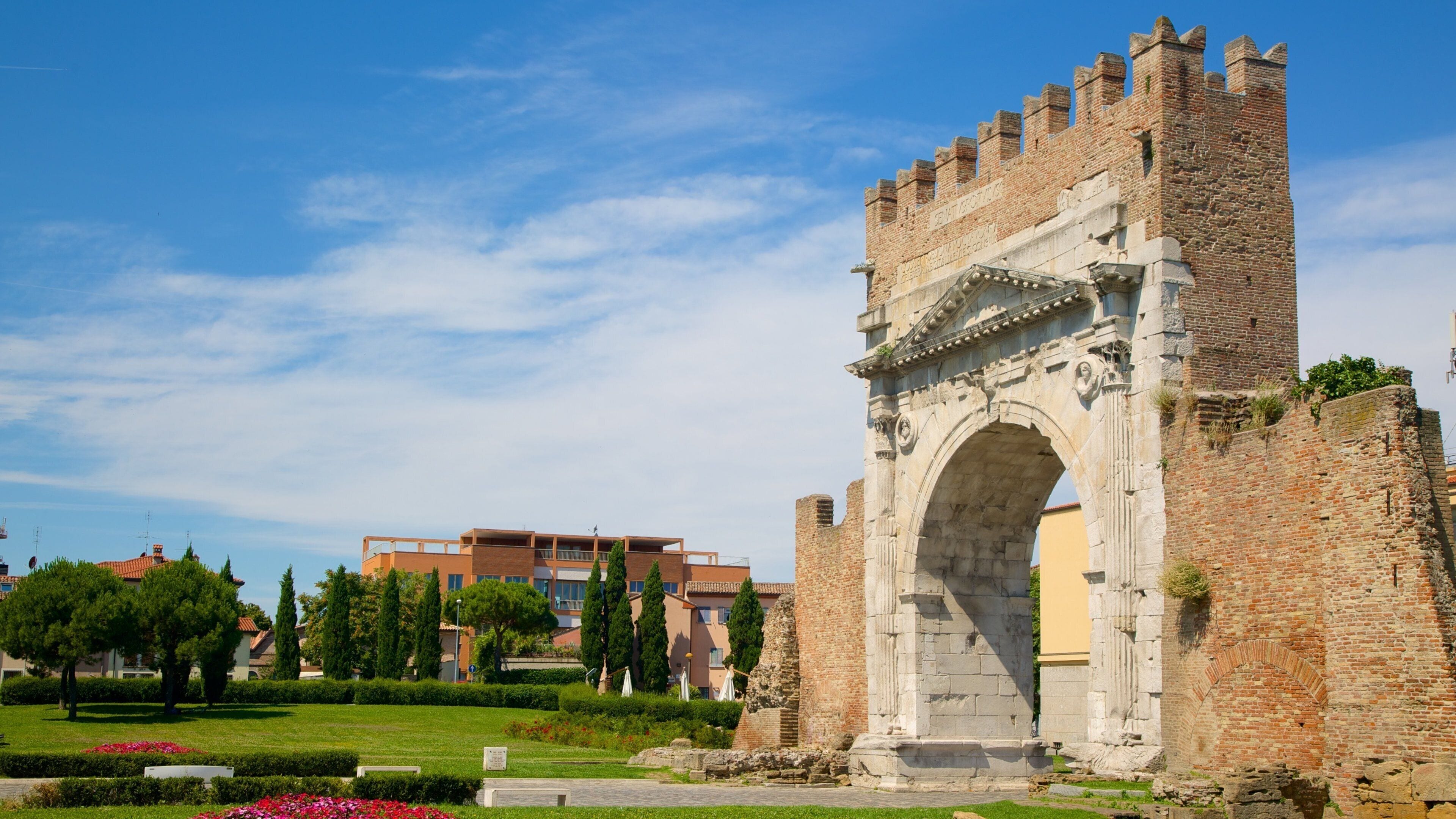 Un arco de piedra histórico con almenas, rodeado de jardines cuidados y edificios modernos.