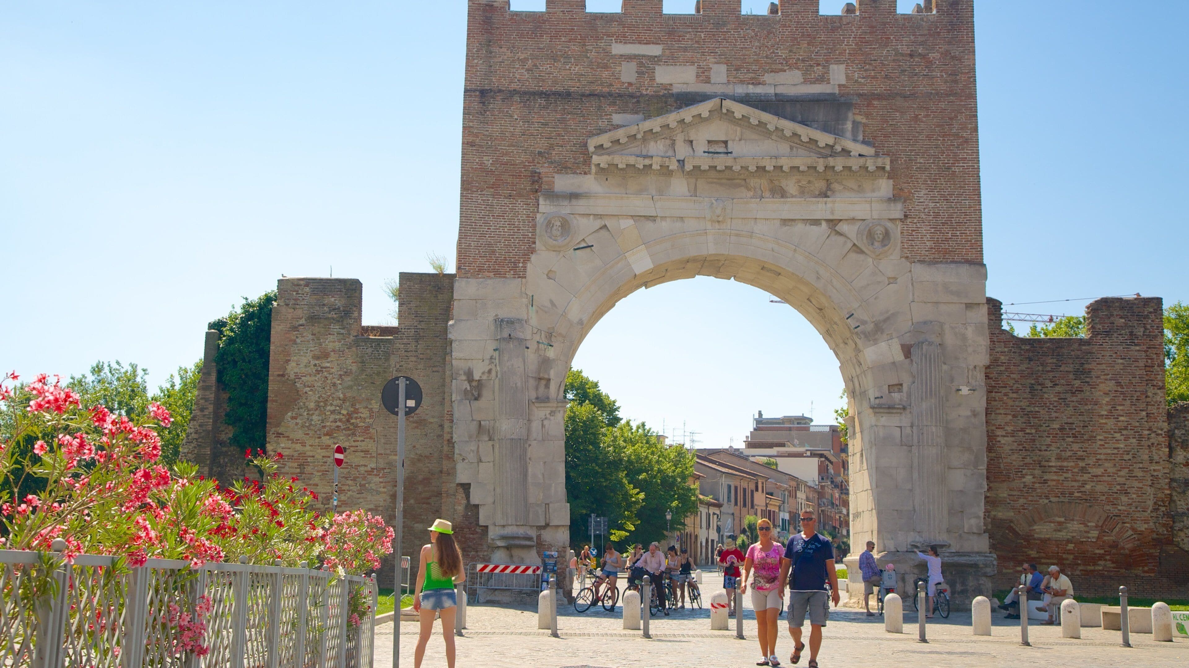 A large stone archway with a brick tower behind it, surrounded by greenery and people walking and cycling.