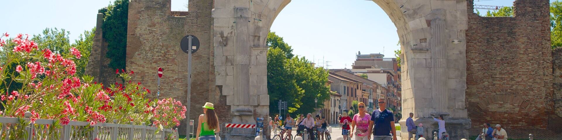 A large stone archway with a brick tower behind it, surrounded by greenery and people walking and cycling.