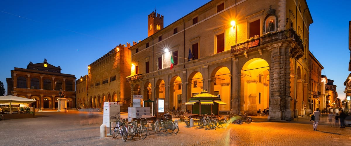 A historic building with arched windows and a flag, illuminated by street lamps at dusk.