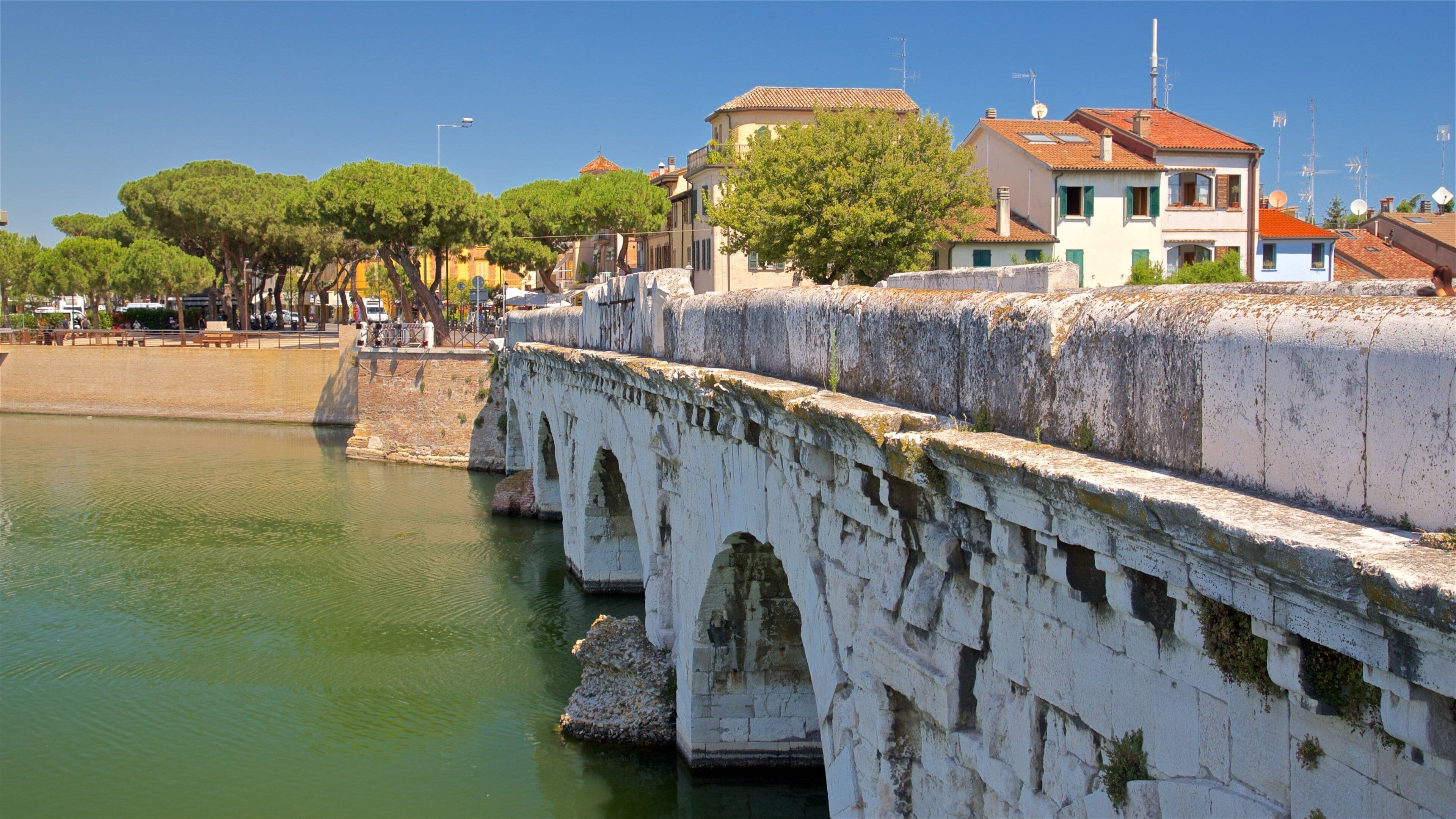 Un puente de piedra sobre un río tranquilo, rodeado de árboles y edificios con tejados rojos.