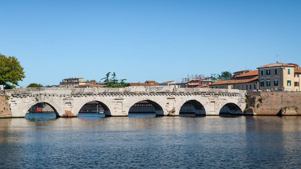 Un pont en pierre, doté de plusieurs arches, enjambe une rivière paisible, et est entouré d’immeubles et d’arbres.