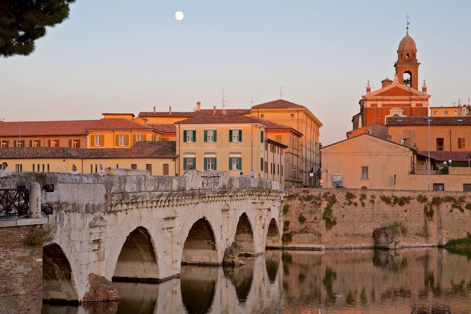 Un pont de pierre enjambe une rivière paisible, et son reflet se mire dans l’eau, où l’on aperçoit les bâtiments environnants ainsi qu’une église surmontée d’un dôme.