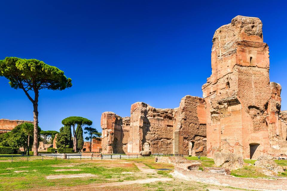 Ancient ruins with a clear blue sky, a large tree, and a well-maintained grassy area.