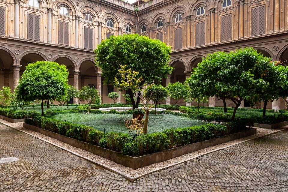 Un cortile con una fontana al centro, circondato da siepi e alberi curati.