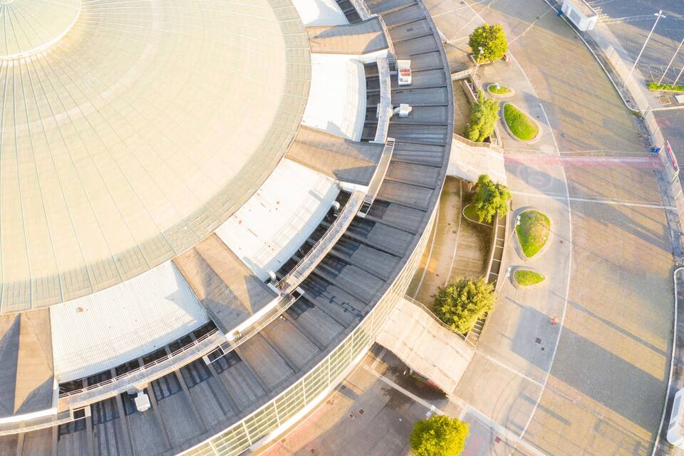 An aerial view of a stadium with a large circular roof, seating areas, and landscaped green spaces.