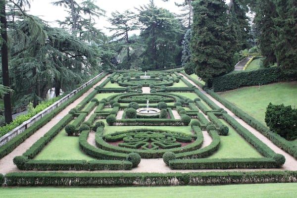 Ein formeller Garten mit geometrischen Hecken, einer zentralen Fontäne und einer Treppe, die zu einer Aussichtsplattform führt.