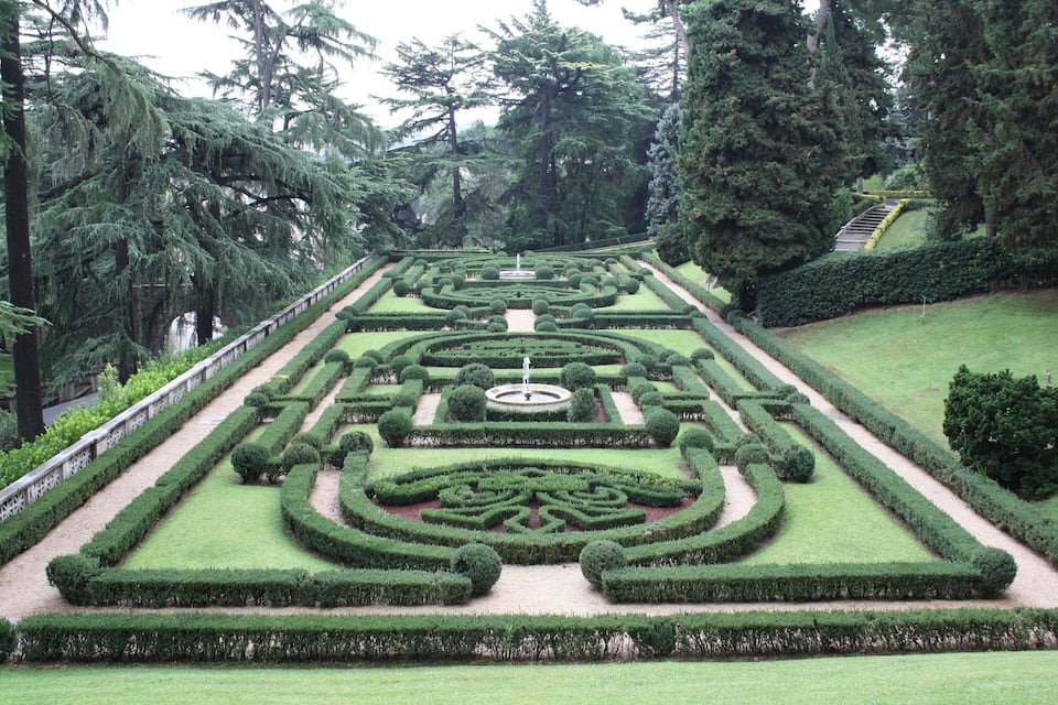 A formal garden with geometric hedges, a central fountain, and a staircase leading up to a viewing platform.