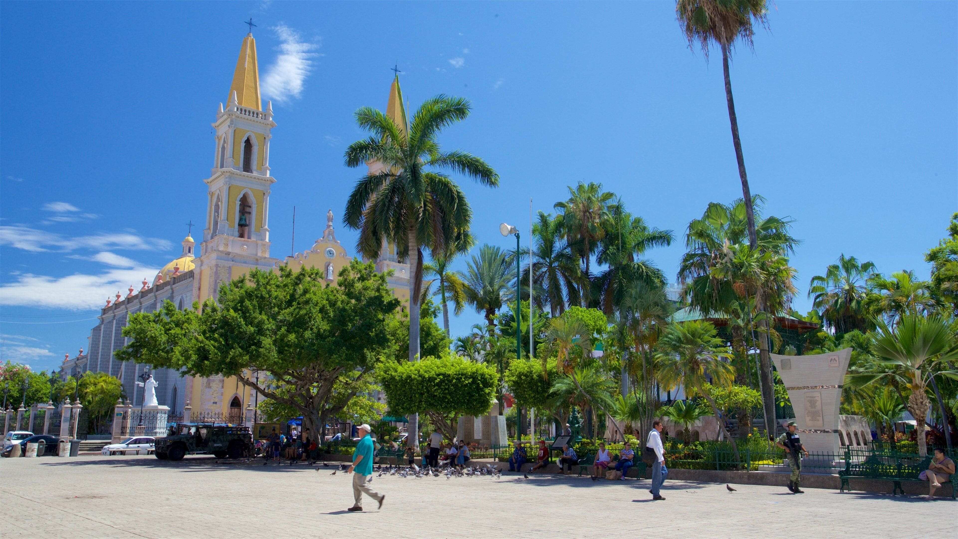 A plaza with palm trees, a church with a tall tower, and people walking.