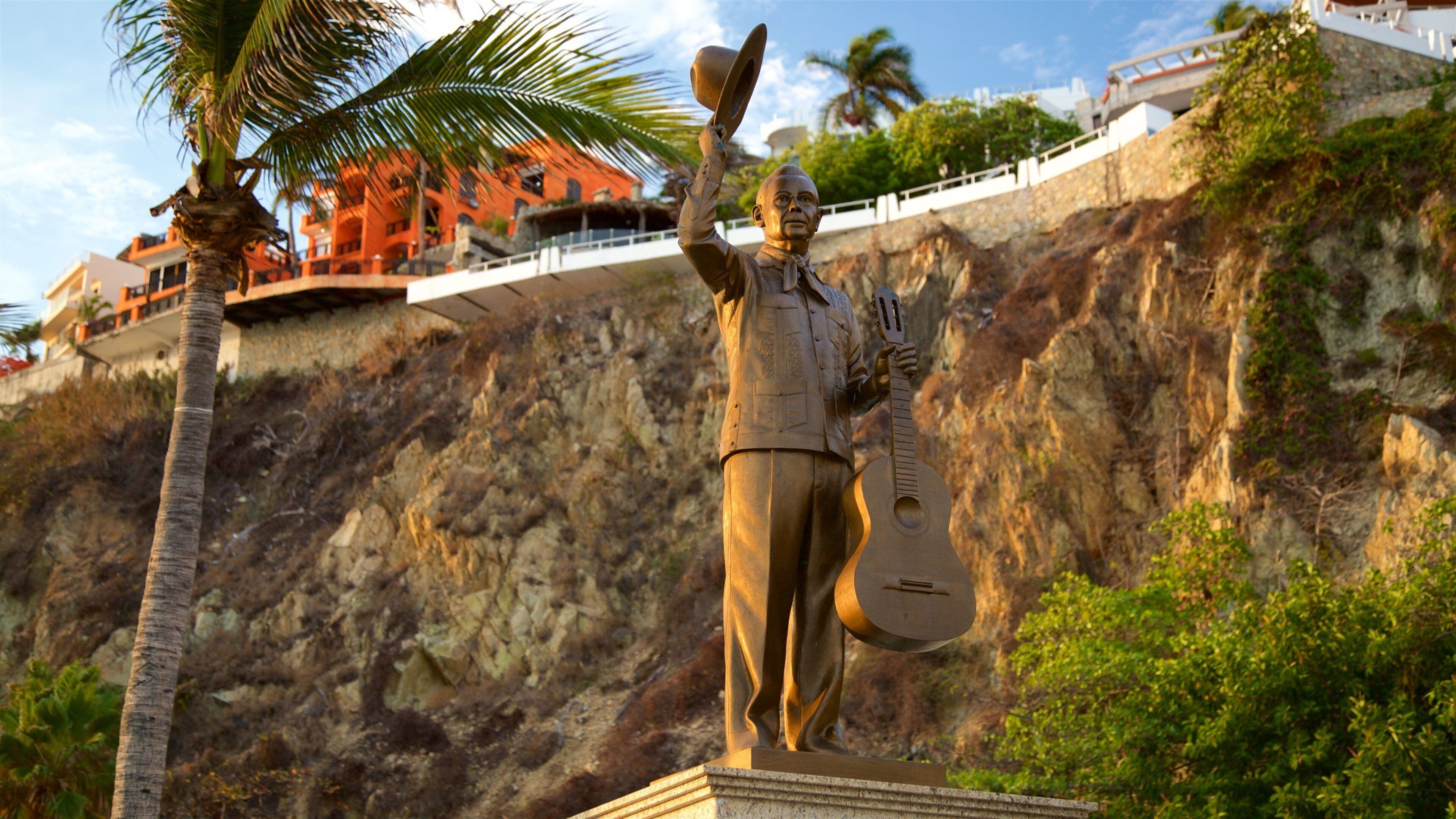 A statue of a man holding a hat and a guitar, with a rocky cliff and a building in the background.
