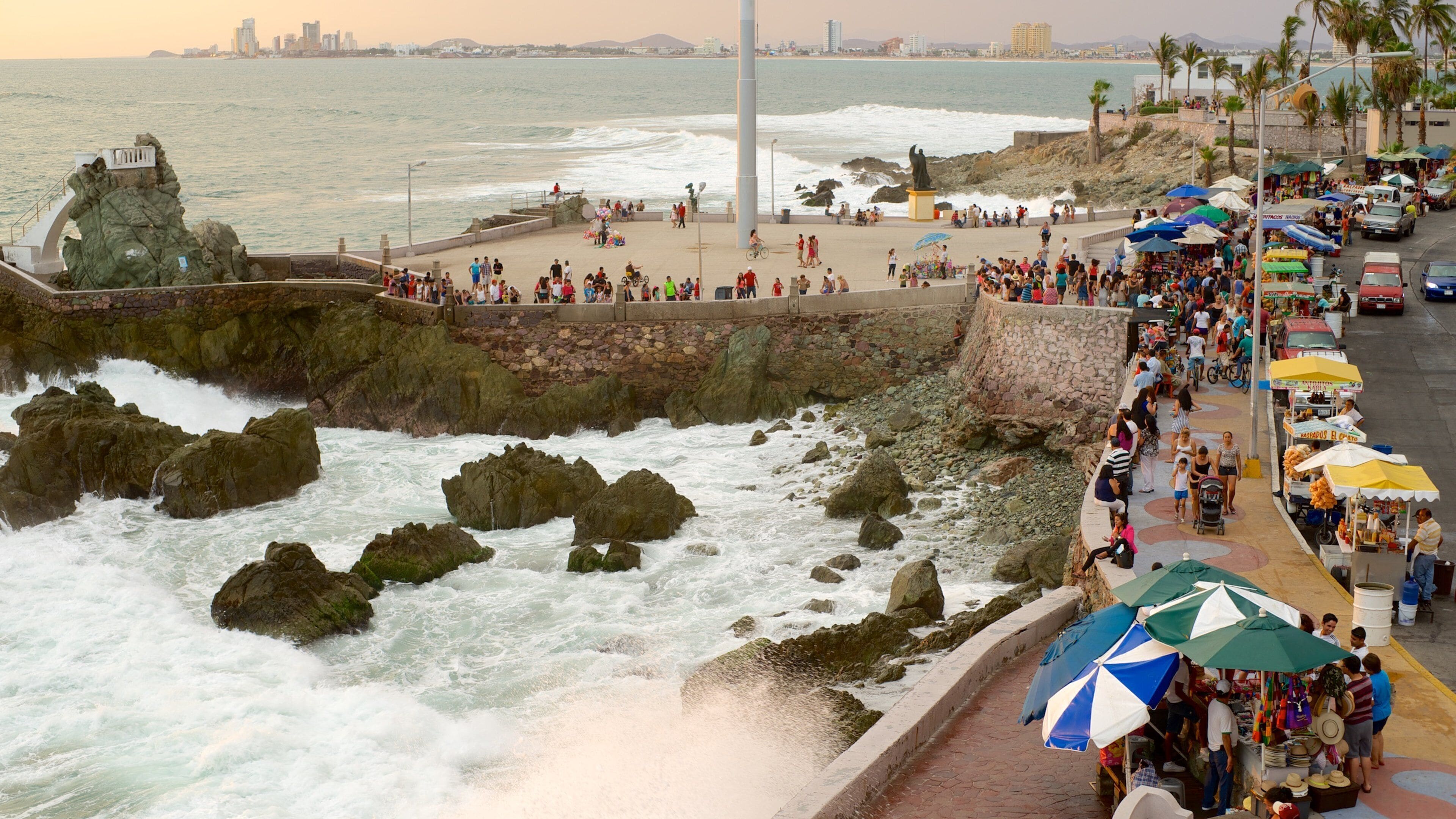 A coastal promenade with a rocky shoreline, a statue, and a bustling market area with vendors and umbrellas.