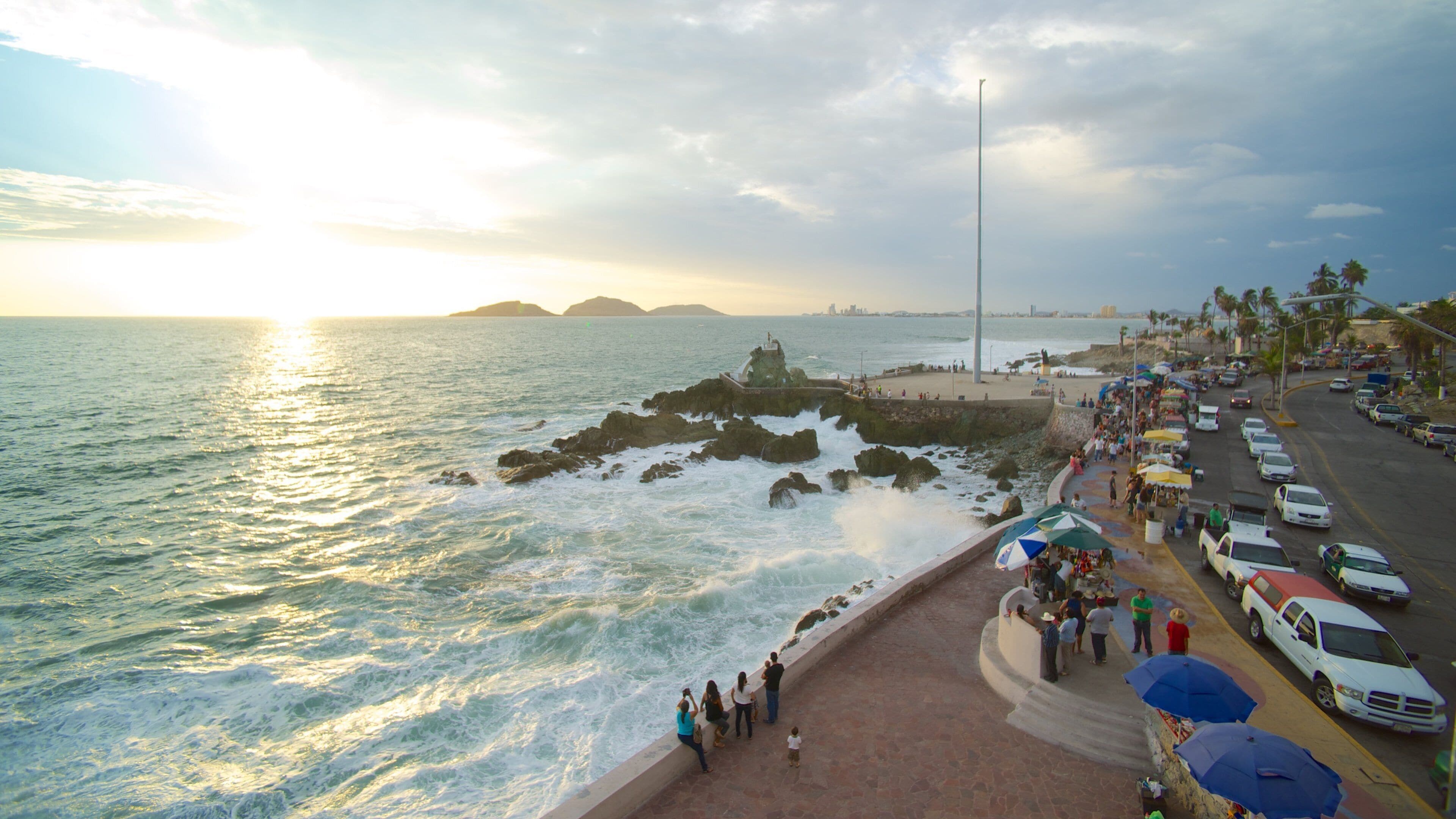 A coastal cityscape with a rocky breakwater, a promenade, parked cars, and a sunset over the ocean.