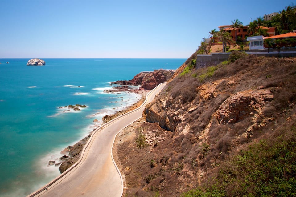 A coastal road with a beach, rocky cliffs, and a building with a red roof.