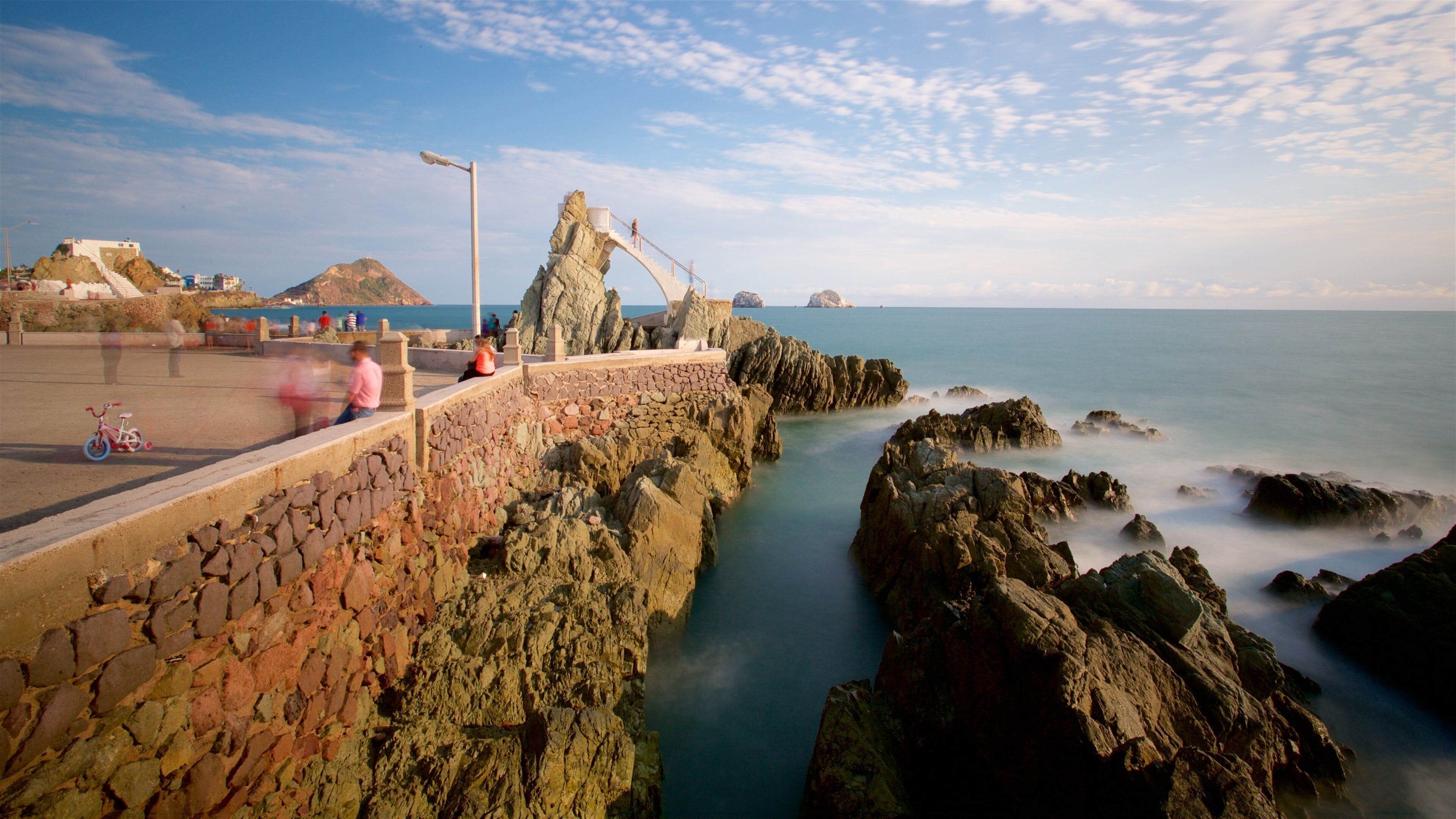 A rocky coastal walkway with a stone wall, a metal street lamp, and a distant view of the sea.