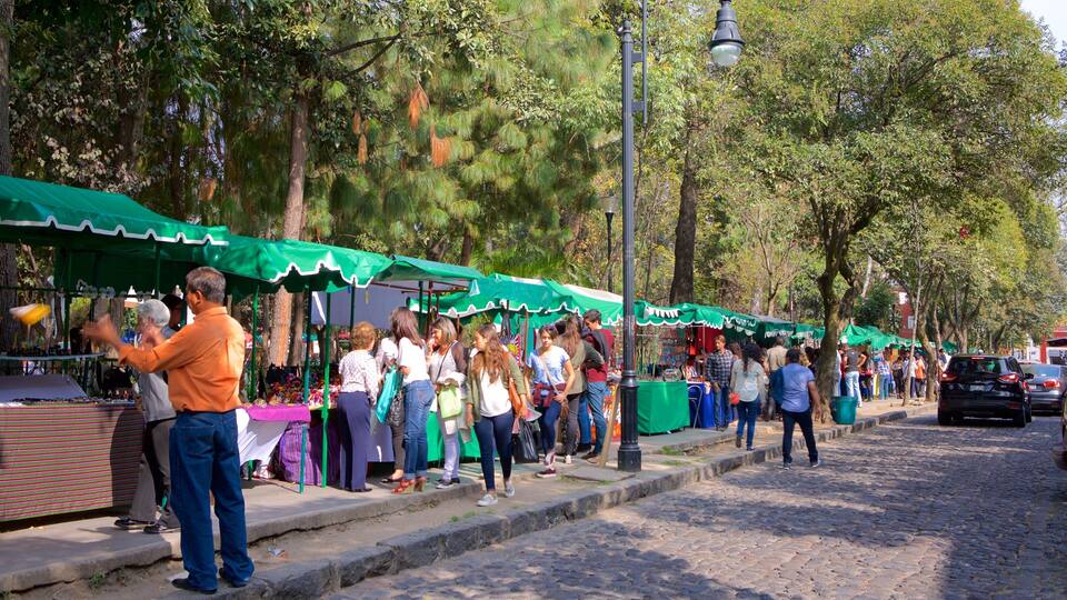A street market with green canopies, people shopping, and trees lining the road.