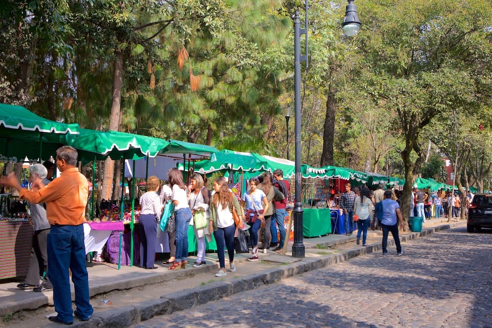 A street market with green canopies, people shopping, and trees lining the road.