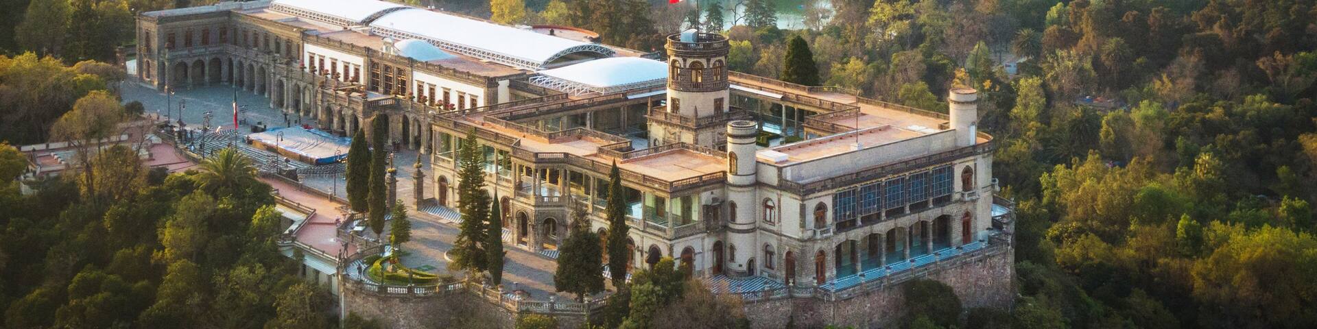 A historic building surrounded by lush greenery, with a cityscape in the background.