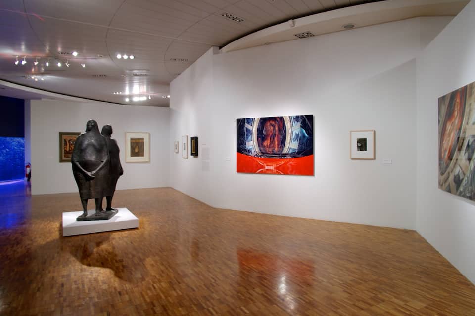 An art gallery with wooden floors, white walls, and various artworks including a large painting with a red and blue background.