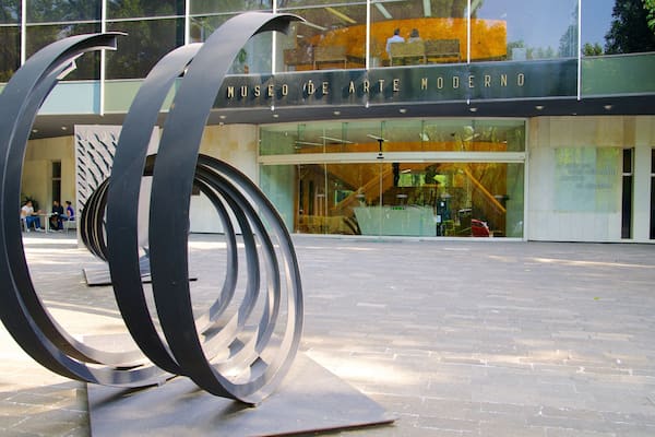 Une sculpture moderne se trouve devant un bâtiment à la façade en verre, sur laquelle on peut lire les mots « Museo de Arte Moderno ».