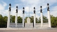 Ein Denkmal mit sechs Säulen, jede bekrönt von einer Statue einer geflügelten Figur, und einer zentralen Statue einer sitzenden Figur.