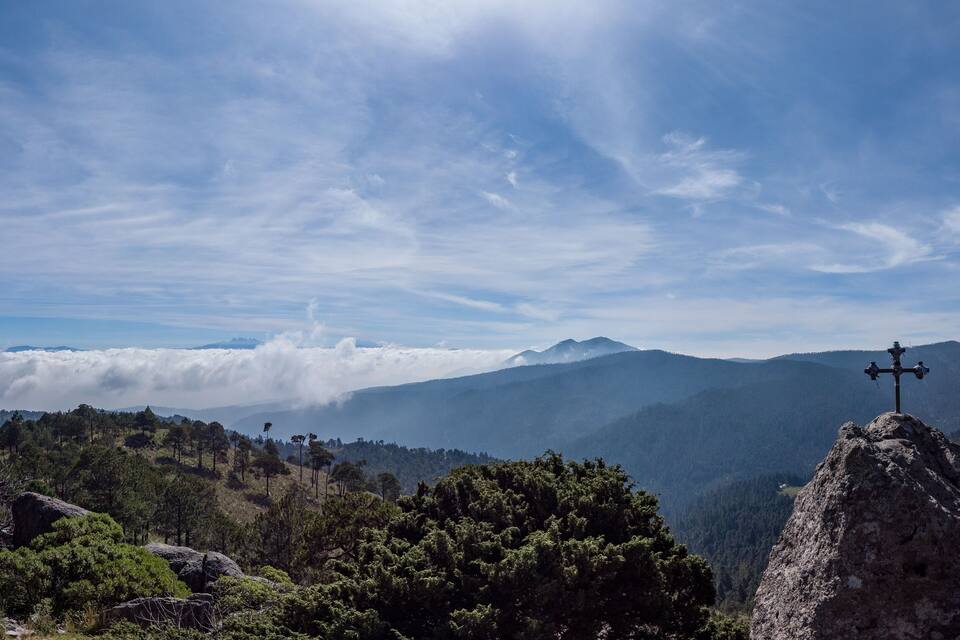 Paisagem de montanha com uma cruz sobre rocha, nuvens abaixo e colinas arborizadas.