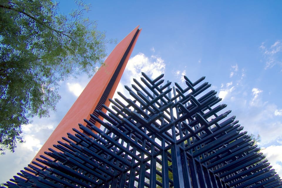 A tall, modern sculpture with a red and black color scheme, set against a clear blue sky with some clouds.