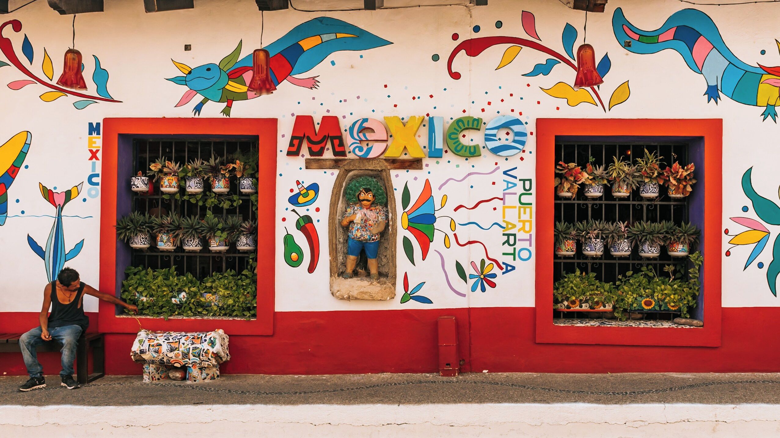 A colorful storefront with large windows displaying potted plants, a mural with vibrant designs, and a person sitting on a bench outside.