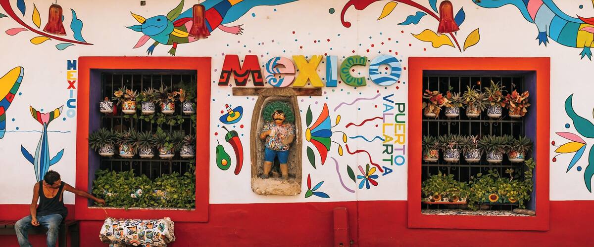 A colorful storefront with large windows displaying potted plants, a mural with vibrant designs, and a person sitting on a bench outside.