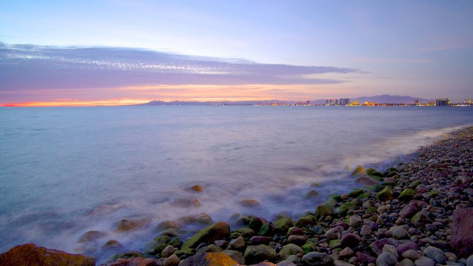 A rocky beach with a calm sea, a sunset, and distant city lights.
