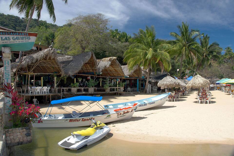 Una playa con chozas de techo de paja, palmeras y barcos.