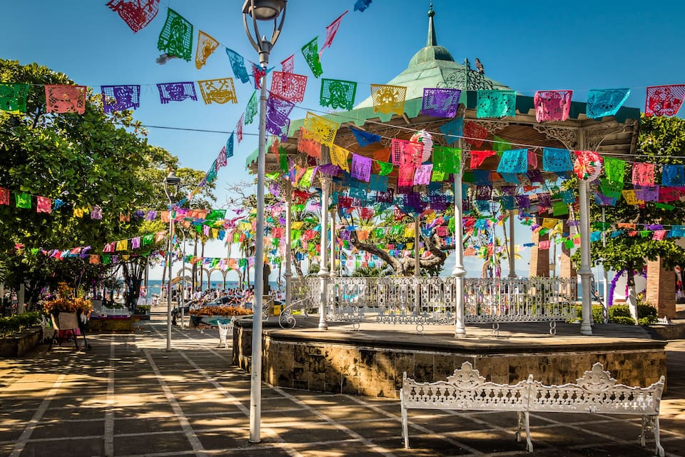 Una plaza con coloridas banderas de papel picado, un pabellón y un banco blanco.