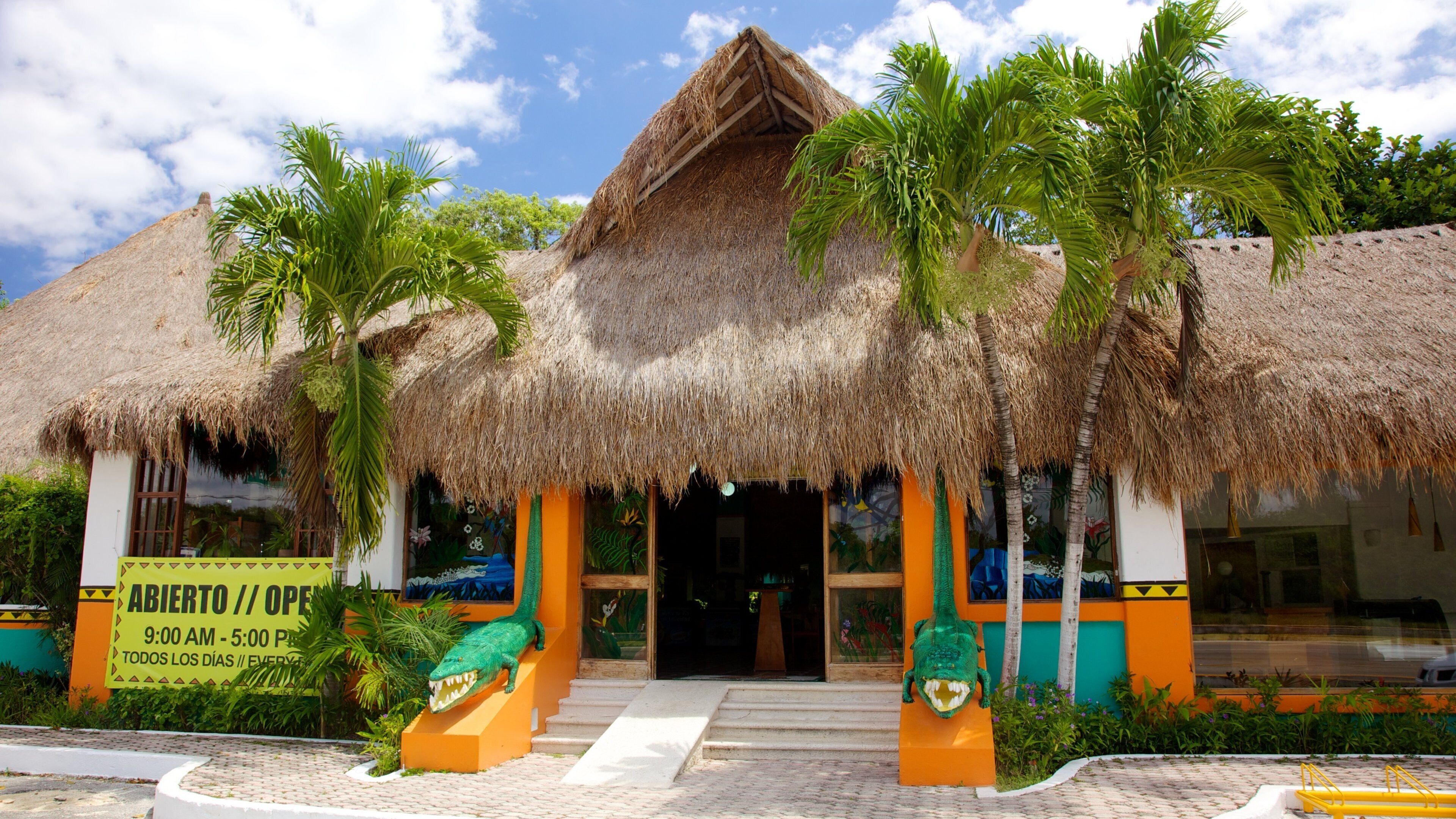 A thatched-roof building with palm trees, a signboard, and a crocodile statue.