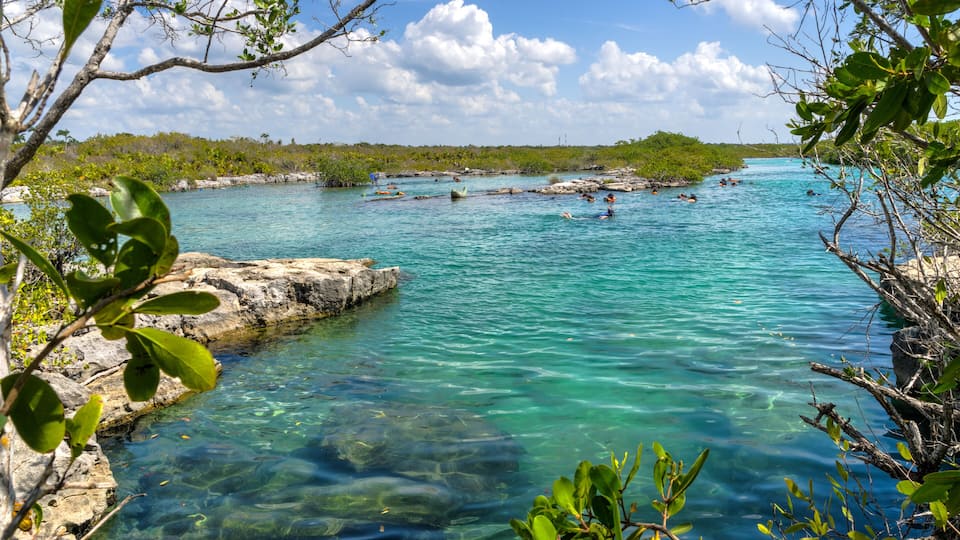 A clear turquoise sea with rocky shores, surrounded by lush greenery and distant land.