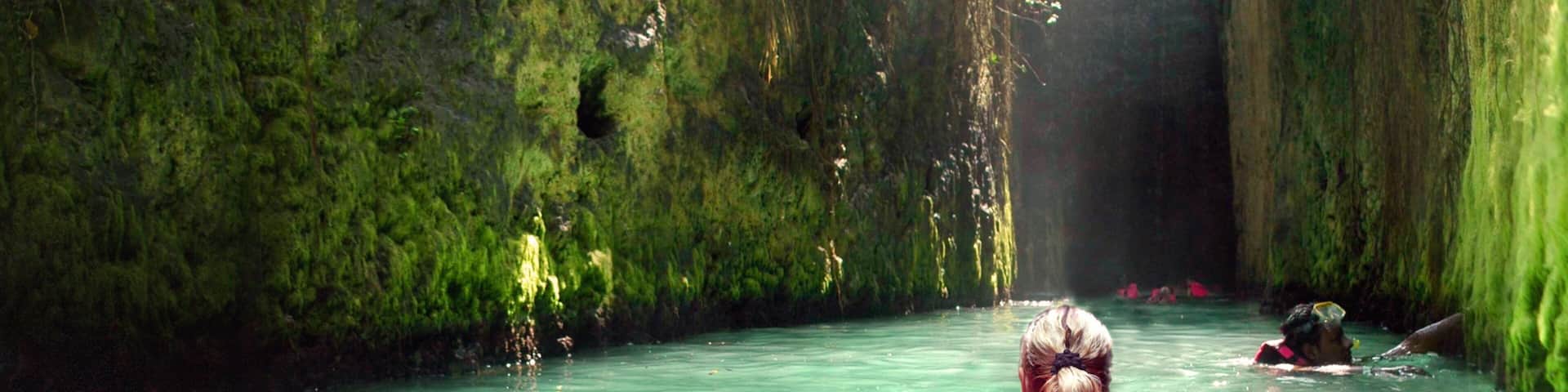 Una persona con traje de baño rosa nada en una piscina turquesa cristalina rodeada de muros verdes cubiertos de musgo.