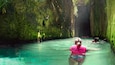 Una persona con traje de baño rosa nada en una piscina turquesa cristalina rodeada de muros verdes cubiertos de musgo.