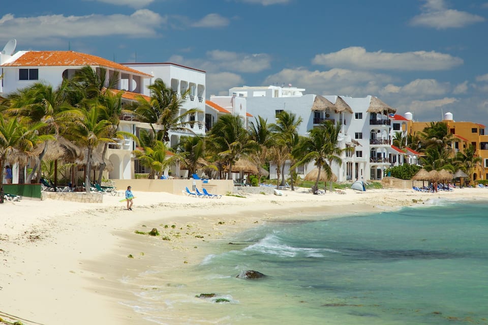 A beach with white buildings, palm trees, and clear blue water.