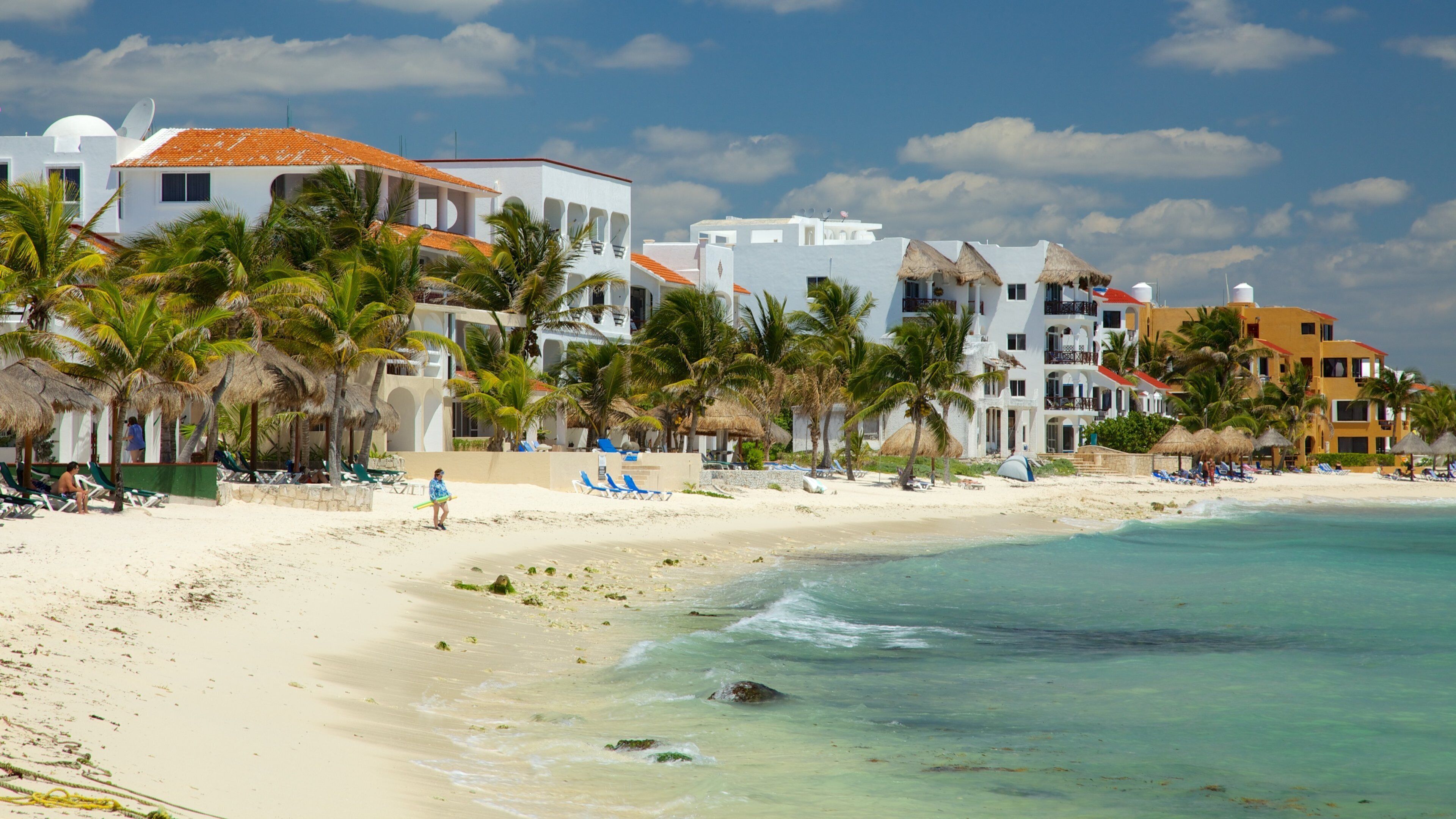 A beach with white buildings, palm trees, and clear blue water.