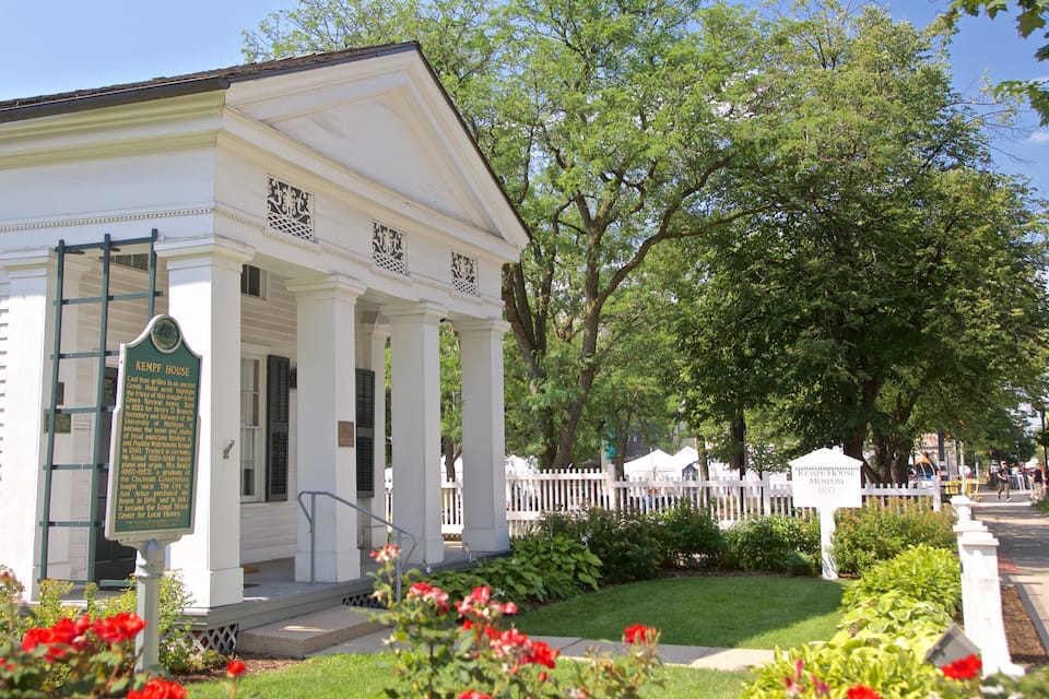 A white historic building with columns and a signboard, surrounded by greenery and a white picket fence.