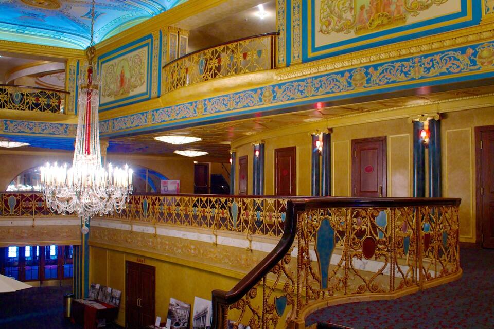 Um interior imponente com grades douradas ornamentadas, um candelabro e detalhes decorativos nas paredes.