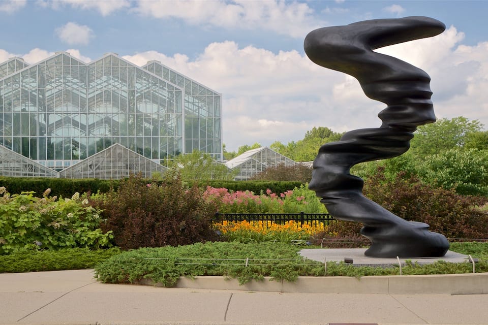 A sculpture in a garden with a greenhouse in the background.