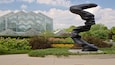 A sculpture in a garden with a greenhouse in the background.