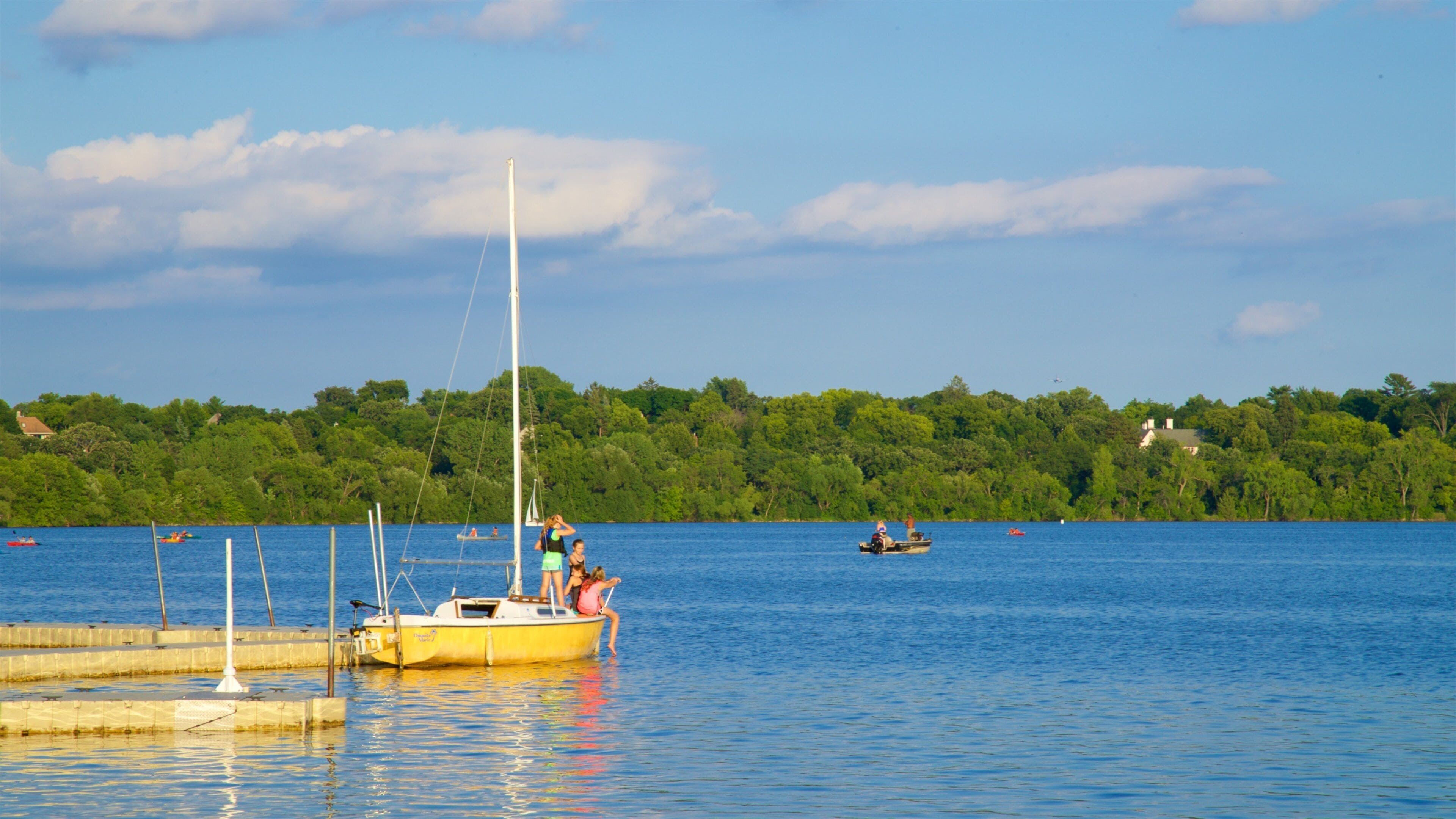 Un lago tranquilo con un velero amarillo amarrado en el muelle, rodeado de frondosos árboles verdes y un cielo azul despejado.