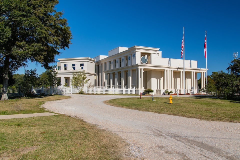 Un edificio grande e histórico con fachada blanca, columnas y dos banderas estadounidenses.