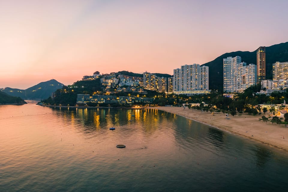 A coastal cityscape at dusk with illuminated buildings, a calm sea, and a mountainous backdrop.