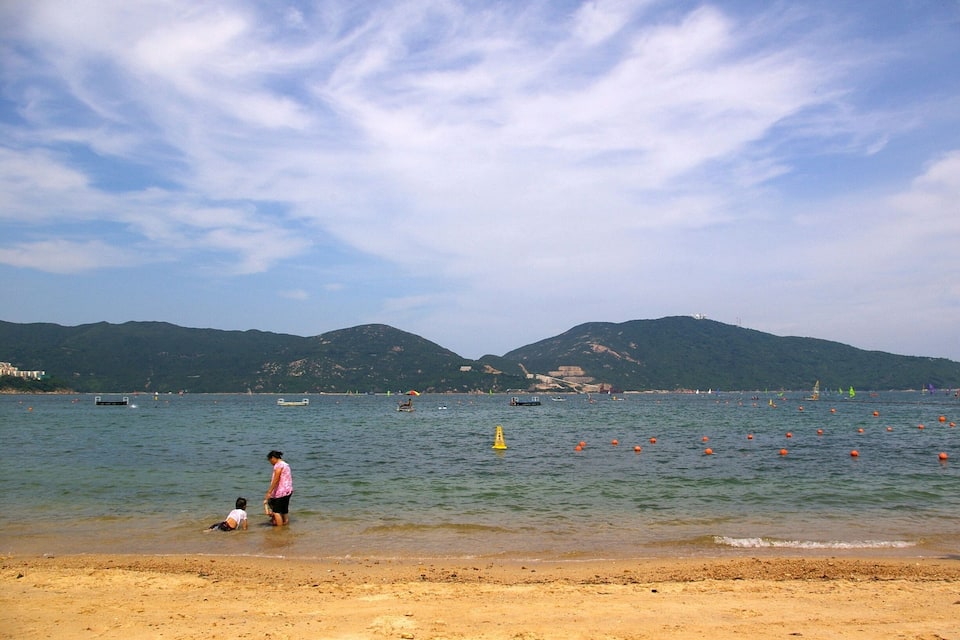 A beach with a woman and a child playing in the water, boats, and mountains in the background.