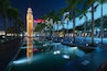 Paisagem urbana noturna com uma torre do relógio imponente, palmeiras ao longo de uma piscina refletora e edifícios modernos ao fundo.