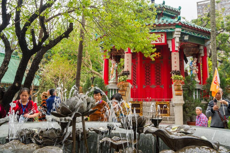 A traditional Chinese pavilion with red walls and green roof, surrounded by trees and a water fountain.